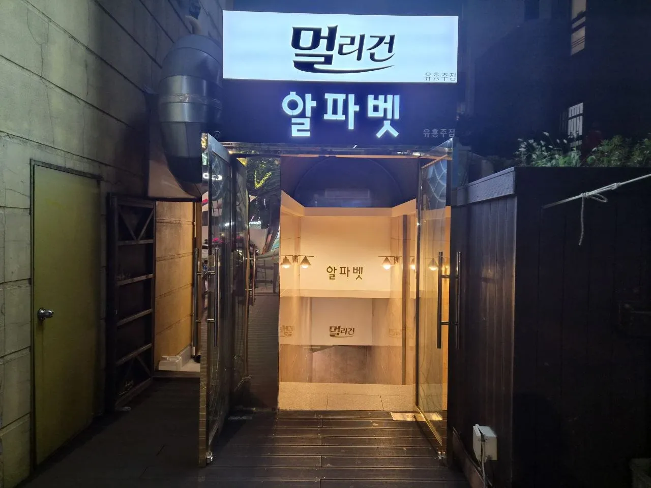 강남 멀리건 간판과 알파벳 간판이 함께 달린 건물 입구. 유리문이 열려 있고 안쪽 계단까지 보인다
